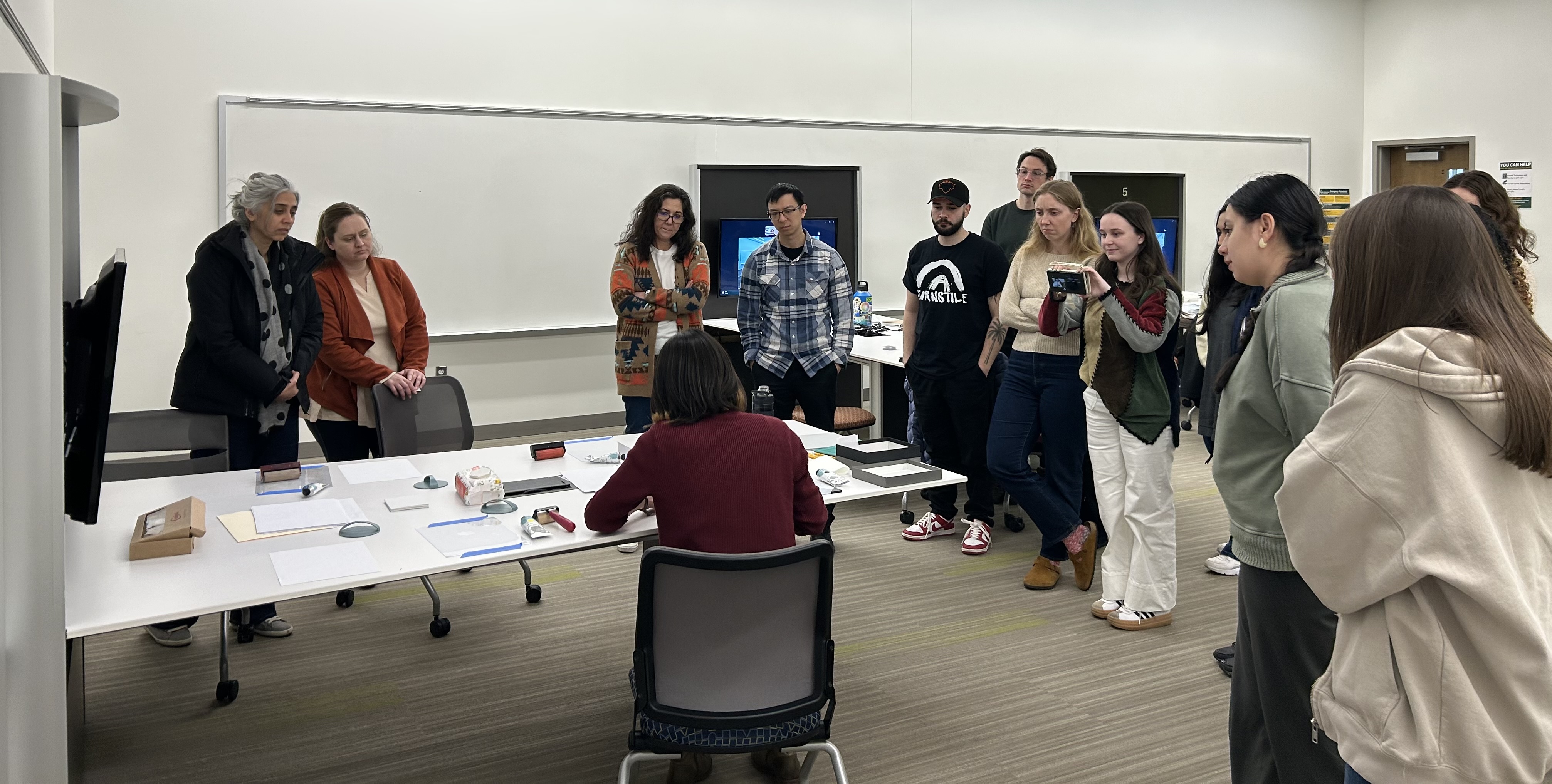 13 students are standing arounda table where the professor is demonstrating printmaking techniques. The professor is sitting and has her back turned from the camera.